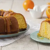 Foto com um bolo redondo de laranja, decorado com calda e raspas de laranja e uma fatia deste mesmo bolo num prato azul do lado direito da foto