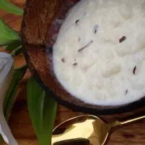 Foto da receita de canjica de coco fresco servida em um pote de vidro sobre uma mesa de madeira com uma colher dourada e um flor branca ao lado
