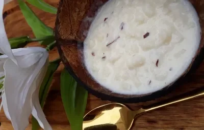 Foto da receita de canjica de coco fresco servida em um pote de vidro sobre uma mesa de madeira com uma colher dourada e um flor branca ao lado