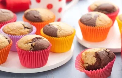 Fotografia em tons de laranja em uma bancada de madeira clara, um prato branco redondo e várias forminhas coloridas, laranja, amarelo, vermelho e roxo com o muffin mesclado de chocolate branco e preto dentro delas.