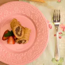 Fotografia em tons de rosa em uma mesa de madeira, uma toalha branca com flores rosas, um prato redondo raso rosa com os rolinhos recheados de chocolate e morango. Ao lado, um garfo.