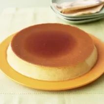 Fotografia de um pudim com calda de caramelo por cima, servido em um prato amarelo sobre uma mesa de madeira