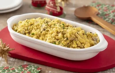 Foto da receita de Farofa Vegana de Legumes. Observa-se um refratário branco com a farofa dentro sobre um guardanapo vermelho. Itens de decoração de Natal decoram a foto.