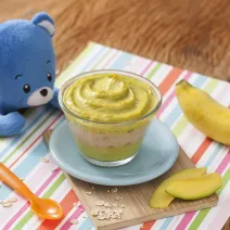 Fotografia em tons de verde em uma bancada de madeira, com um paninho listrado colorido e uma tacinha transparente com abacate e banana apoiado em um pratinho azul. Ao lado, um ursinho de pelúcia azul, uma colher laranja e uma banana.