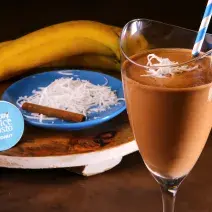 Fotografia em tons de azul em uma bancada de madeira, uma tábua de madeira com um copo de vidro alto e a bebida de chocolate com coco e banana dentro dele. Ao fundo, um prato azul com raspas de coco, uma canela em pau e uma banana.