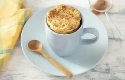 Fotografia em tons de azul em uma bancada de madeira clara, um prato e uma xícara azul ao centro com o bolo de caneca dentro. Ao lado, um pano amarelo e uma peneirinha com canela.