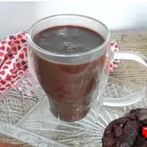 Foto da receita de Chocolate Quente. Observa-se uma xícara branca transparente com a bebida dentro, sobre um pires transparente de vidro.