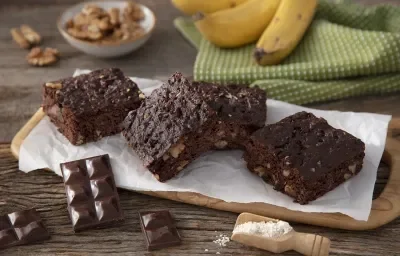 Fotografia em tons de marrom em uma bancada de madeira de cor marrom. Ao centro, uma tábua de madeira contendo os brownies em cima. Ao lado, há alguns tabletes de chocolate e ao fundo, um pano verde quadriculado com algumas bananas em cima. Fotografia em tons de marrom em uma bancada de madeira de cor marrom. Ao centro, uma tábua de madeira contendo os brownies em cima. Ao lado, há alguns tabletes de chocolate e ao fundo, um pano verde quadriculado com algumas bananas em cima.