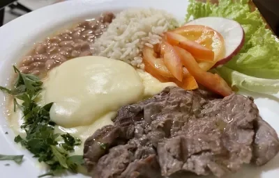 Foto da receita de Purê Francês. Observa-se um prato com um cremoso purê de batatas, acompanhado de arroz, feijão, iscas de carne e uma salada de tomate, rabanete e alface crespa.