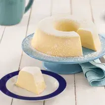 Fotografia em tons de azul em uma bancada de madeira branca, um suporte para bolo azul claro com o pudim de leite cortado ao meio em cima dele. Ao lado, um prato com uma fatia do pudim.