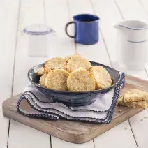 Fotografia em tons de azul em uma bancada de madeira branca, uma tábua de madeira, um paninho branco com detalhes azuis, um recipiente azul redondo fundo com os biscoitos de coco com amêndoas dentro. Ao lado, uma xícara azul.