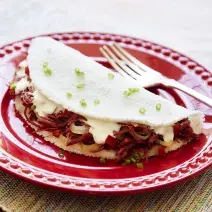 Fotografia em tons de branco e vermelho, com prato vermelho ao centro contendo uma tapioca com recheio de carne-seca, queijo de coalho e salpicada de cebolinha, acompanhada de um garfo, sobre guardanapo multicolorido.