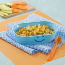 Fotografia em tons de laranja em uma bancada de madeira de cor azul. Ao centro, um pano laranja e um pano quadriculado azul, em cima, há um recipiente contendo os legumes e ao lado, uma colher laranja. Ao fundo, uma tábua contendo alguns legumes.