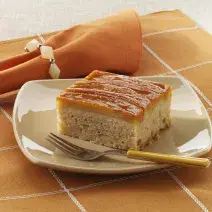 Foto de um prato branco em cima de uma toalha laranja com branco e com um pedaço de bolo de banana dentro com um garfo do lado