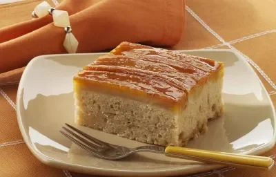 Foto de um prato branco em cima de uma toalha laranja com branco e com um pedaço de bolo de banana dentro com um garfo do lado