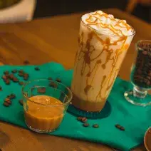 Fotografia em tons de verde de uma mesa de madeira com um paninho verde e ao centro um copo decorado com doce de leite, com uma bebida de leite e decorado com chantilly. Ao redor cápsula e grãos de café.