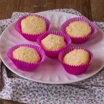 Imagem da receita de Queijadinha da Miriam Braga, servida em forminhas rosas de cupcakes, sobre um prato rosa, em um bancada de madeira decorada com um tecido florido