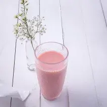 Fotografia em tons de rosa em uma bancada de madeira clara com um copo de vidro ao centro e a vitamina de hortelã com goiaba dentro dele. Ao fundo, um vasinho de vidro com flores brancas pequenas.