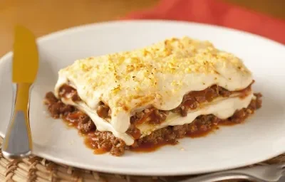 Foto de uma fatia de lasanha em um prato branco com um garfo e uma faca do lado