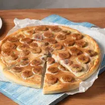 Fotografia em tons de bege e azul, de uma bancada de madeira, ao centro a pizza com rodelas de banana é servida em um papel de seda e uma tábua azul. Ao fundo canela e pratos brancos para decorar.