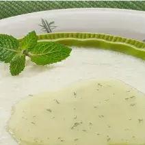 Fotografia em tons de verde em uma mesa com uma toalha verde, um prato branco grande redondo, outro prato branco redondo fundo verde com a mousse de pera com a calda de hortelã.