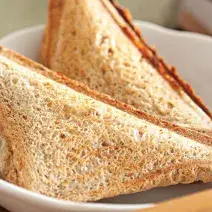 Fotografia em tons de bege em uma mesa com uma toalha bege, um prato redondo fundo branco com dois sanduíches de pão integral tostados e recheados com queijo e lombo.