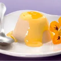 foto em tons lilas e branco. Acima de um prato roxo contém um prato na cor branco que comporta Flan. Ao lado uma colher e uma flor para decorar.o