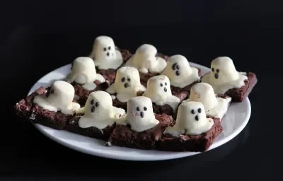 Fotografia de um prato branco com pedaços de brownie de chocolate cortados em quadrados, com marshmallows derretidos em cima representando um fantasma. Fotografia de um prato branco com pedaços de brownie de chocolate cortados em quadrados, com marshmallows derretidos em cima representando um fantasma.