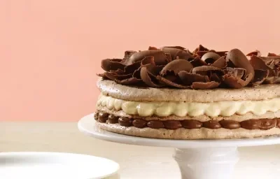 Fotografia em tons de rosa em uma mesa de madeira branca com um pano de prato vermelho, uma parede de fundo rosa, um suporte para bolo branco com a torta de macaron de chocolate e creme de avelã.