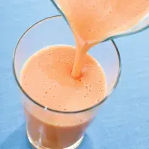 fotografia em tons de azul e laranja vista de uma bancada azul, contém um copo transparente com a vitamina e uma jarra despejando a vitamina no copo