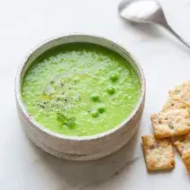 Fotografia em tons de verde em uma bancada de madeira de cor branca. Ao centro, uma tigela contendo a sopa de ervilha com alguns biscoitos de água e sal e uma colher ao lado.