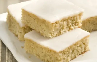 Fotografia em tons de bege em uma mesa de madeira com uma toalha bege listrada, um prato branco quadrado com pedaços de bolo de camomila com cobertura de glacê branco.