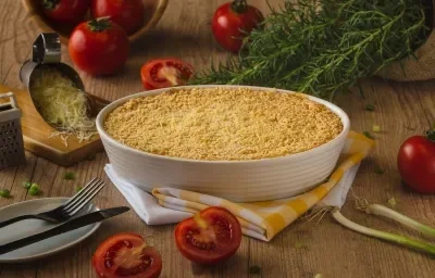 Fotografia em tons de amarelo em uma bancada de madeira escura, um recipiente branco oval com o escondidinho de carne suína gratinado dentro dele, um pano amarelo xadrez e alguns elementos para compor a foto, como tomate, queijo ralado e alecrim.