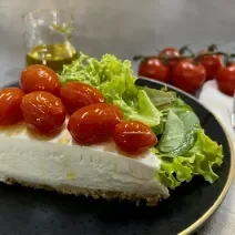 Foto da receita de Cheesecake Salgado. Observa-se uma fatia da torta com tomatinhos confit em cima em um prato preto servido com uma salada de alface.