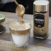 Fotografia em tons de marrom de uma bancada de madeira, sobre ela um copo de vidro com o dalgona coffé e uma colher. Ao lado um recipiente do Nescafé gold.