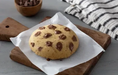 Fotografia em tons de marrom em uma bancada de madeira de cor cinza. Ao centro, uma tábua de madeira com um pedaço de papel manteiga em cima contendo o cookie. A fundo, há um pote com pedaços de chocolate e um pano branco listrado.