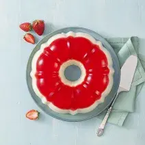 Fotografia em tons de cinza e vermelho de uma bancada cinza vista de cima, um prato azul redondo contém a gelatina nas cores vermelha e branca, ao lado um pano azul e uma espátula por cima, e do outro lado pedaços de morangos.