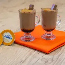 Fotografia em tons de laranja em uma bancada de madeira de cor marrom. Ao centro, um pano laranja contendo duas taças com as bebidas. Ao lado das taças há duas cápsulas de Nescafé.