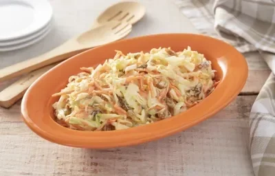 Fotografia em tons de laranja em uma bancada de madeira clara, um recipiente oval laranja com a salada de repolho, cenoura, uvas-passas e maionese. Ao lado, colheres de pau e um paninho bege xadrez. Fotografia em tons de laranja em uma bancada de madeira clara, um recipiente oval laranja com a salada de repolho, cenoura, uvas-passas e maionese. Ao lado, colheres de pau e um paninho bege xadrez.