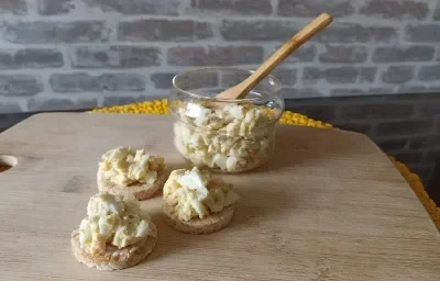 Imagem da receita de Patê de Ovo, em uma bolacha, sobre uma tábua e ao lado uma colher de madeira em um pote de vidro