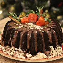 Fotografia em tons de marrom e vermelho de uma bancada de madeira clara, sobre ela um prato vermelho redondo com o bolo de chocolate decorado. Ao lado uma bolinha natalina e uma pinha.