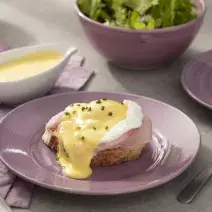 Fotografia em tons de lilás em uma bancada de madeira clara, um paninho lilás, um prato redondo lilás com o ovo benedict em cima dele, com a fatia de pão tostado, presunto, o ovo e cebolinha-verde para decorar. Ao fundo, um bowl com salada verde.