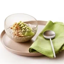 Foto da receita de Overnight Oats de Kiwi. Observa-se um prato de cerâmica marrom com um recipiente de vidro em cima com a receita. Ao lado direito, um guardanapo verde com uma colherinha.