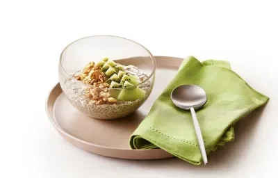 Foto da receita de Overnight Oats de Kiwi. Observa-se um prato de cerâmica marrom com um recipiente de vidro em cima com a receita. Ao lado direito, um guardanapo verde com uma colherinha.