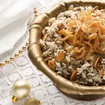 Fotografia em tons de marrom e dourado em uma mesa de madeira com uma toalha branca de renda com um recipiente redondo fundo e grande dourado com o arroz de festa com lentilhas.