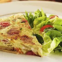 Fotografia em tons de verde em uma bancada com um prato branco redondo raso, um pano amarelo de fundo e a tortilla de omelete com batata e tomate em cima dele, ao lado, salada de alface e tomate cereja.