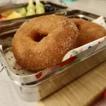 Fotografia em tons de vermelho em uma bancada de madeira clara, uma marmita ao meio com dois donuts de maçã dentro. Ao lado, brinquedos infantis.