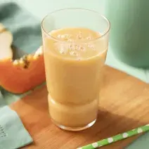 Fotografia em tons de verde e laranja de uma bancada verde, paninho verde, fatia de pera e mamão e uma tábua com um copo com a bebida laranja. Ao fundo uma jarra verde.