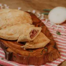 Fotografia em tons de marrom e vermelho de uma bancada de madeira com um paninho listrado vermelho e branco, sobre ele uma tabua de madeira com mini empadas. Ao fundo uma cebola cortada ao meio.