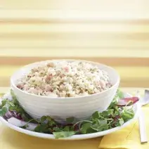 Fotografia em tons de amarelo em uma bancada de madeira clara, um pano amarelo, um prato redondo com salada de alface e cebola roxa e um potinho branco com a salada de arroz dentro dele.